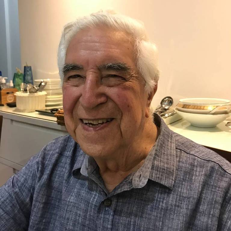 Um homem idoso com cabelo branco e um sorriso amigável. Ele está usando uma camisa azul com padrões. Ao fundo, há uma mesa com utensílios e pratos, sugerindo um ambiente domiciliar