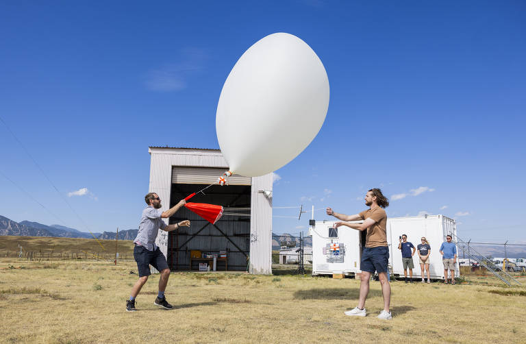 Duas pessoas estão segurando um grande balão branco em um campo aberto, próximo a um galpão. Uma delas está usando uma camisa de manga curta e shorts, enquanto a outra está de camiseta e shorts. Ao fundo, há um grupo de pessoas observando a cena. O céu está claro e azul, e as montanhas são visíveis ao longe.