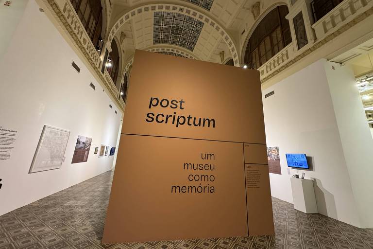 A imagem mostra uma instalação de exposição em um museu, com um grande painel em destaque que diz 'post scriptum' em letras grandes e pretas. Abaixo, está escrito 'um museu como memória'. O ambiente é amplo, com um teto alto e detalhes arquitetônicos visíveis. À esquerda, há uma parede branca com várias obras de arte emolduradas. O piso é decorado com azulejos em padrão geométrico.