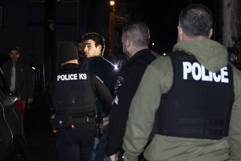 A imagem mostra um grupo de policiais em uma operação noturna. Eles estão vestidos com coletes pretos que têm a palavra 'POLICE' em letras brancas. A cena ocorre em uma rua, com um veículo escuro à esquerda e uma iluminação fraca ao fundo. Os policiais parecem estar em ação, com alguns se movendo em direção a um indivíduo que está sendo abordado.