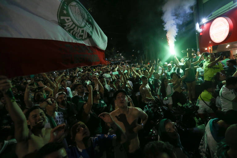 Foto mostra torcida do Palmeiras celebrando título de bicampeão brasileiro, em 2023, no entorno do Allianz Parque, durante a noite. Na imagem é possível ver diversas pessoas com as mãos erguidas. na lateral direita da foto, um dos torcedores segura um sinalizador com a cor verde. Já na ponta esquerda da imagem, outros palestrinos ostentam um bandeirão com o símbolo do clube.