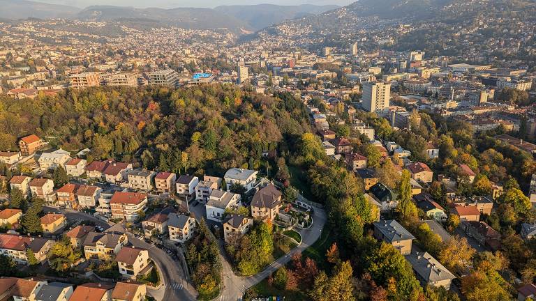 A imagem mostra uma vista aérea de uma cidade situada em uma área montanhosa. No primeiro plano, há várias casas com telhados vermelhos e algumas áreas verdes. Ao fundo, a cidade se estende com prédios e montanhas ao redor, enquanto a vegetação apresenta cores de outono, como amarelo e laranja. A paisagem é marcada por ruas sinuosas e uma mistura de áreas urbanas e naturais.