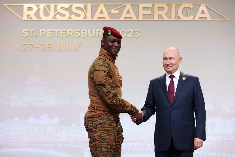 Na imagem, Vladimir Putin e Ibrahim Traore estão se cumprimentando em um evento intitulado 'Rússia-África' que ocorre em São Petersburgo em 2023. Traore está vestida com um uniforme militar camuflado, enquantoPutin usa um terno escuro e uma gravata. Ao fundo, há um painel com o texto 'RUSSIA-AFRICA' e a data do evento.