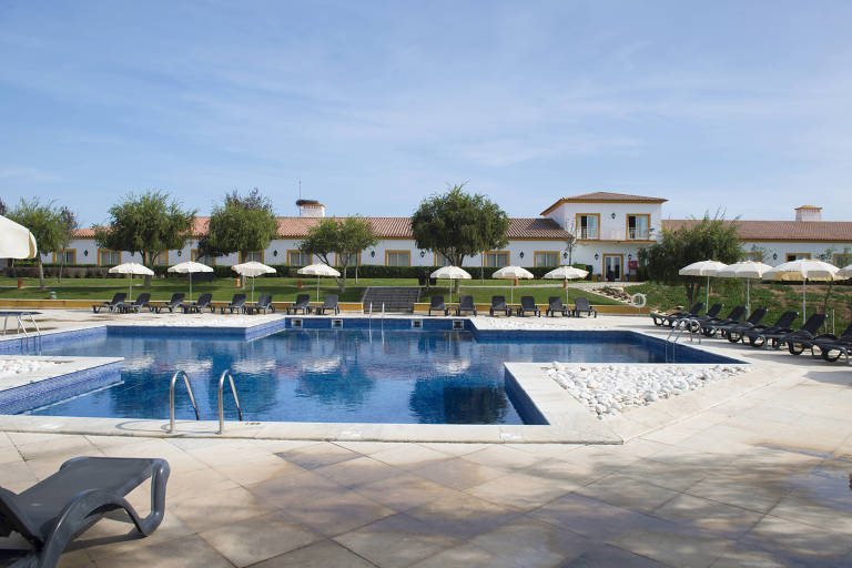 A imagem mostra uma área de piscina em um resort. A piscina é grande e possui água azul clara, com escadas de acesso. Ao redor da piscina, há espreguiçadeiras pretas e guarda-sóis brancos. Ao fundo, vê-se um edifício de dois andares com janelas e um telhado de cerâmica. O ambiente é ensolarado, com céu azul e algumas árvores ao redor.