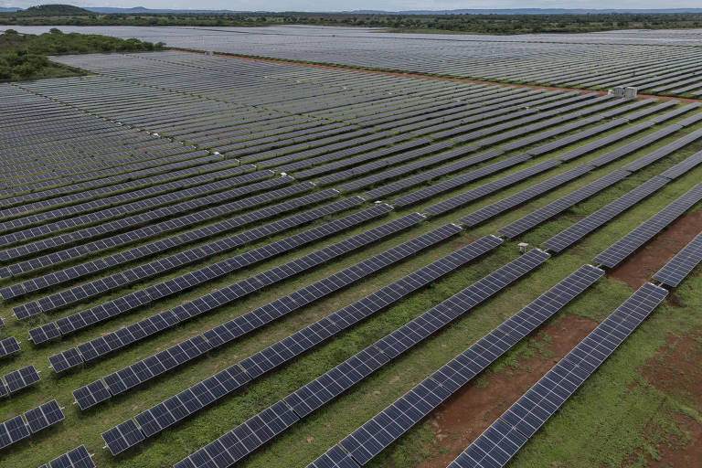 A imagem mostra uma vasta área coberta por painéis solares dispostos em fileiras organizadas. O campo se estende até o horizonte, com uma paisagem verde ao redor e algumas colinas ao fundo. O céu está parcialmente nublado.