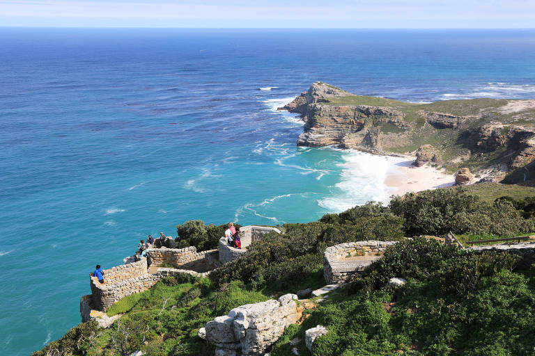 A imagem mostra uma vista panorâmica do Cabo da Boa Esperança, com o oceano azul e calmo à esquerda e uma costa rochosa à direita. Há vegetação verde na encosta e algumas pessoas caminhando em uma trilha. O céu está claro e ensolarado, com poucas nuvens.