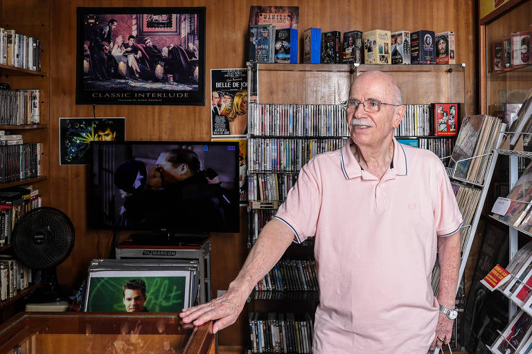 Um homem idoso, com cabelo grisalho e óculos, está em uma loja cheia de CDs e DVDs. Ele usa uma camisa polo rosa e está posicionado ao lado de uma vitrine de madeira. Ao fundo, há prateleiras repletas de CDs e DVDs, além de um televisor exibindo uma cena de um filme. Na parede, há quadros e pôsteres decorativos.
