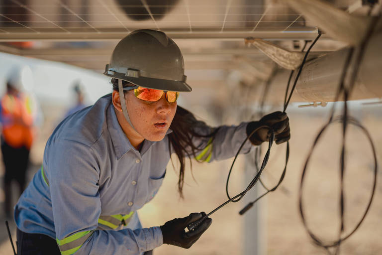 Uma mulher usando um capacete e óculos de proteção está trabalhando sob um painel solar, manipulando fios. Ela veste uma camisa de manga longa com faixas refletivas e parece concentrada na tarefa. Ao fundo, outras pessoas estão visíveis, mas desfocadas.