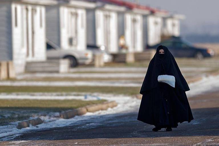 A imagem mostra uma mulher vestindo um manto preto, que deixa à vista apenas seus olhos, caminhando em uma rua de uma área residencial. Ao fundo, há várias casas com telhados vermelhos. O ambiente parece ser frio, com algumas áreas cobertas de neve.
