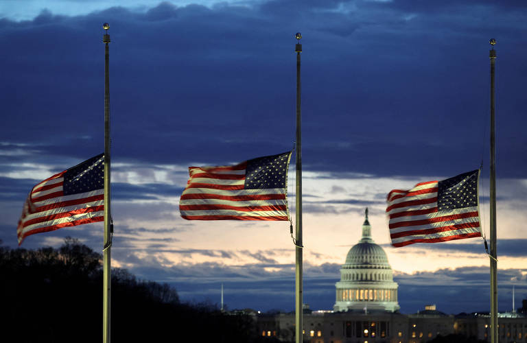 A imagem mostra três bandeiras dos Estados Unidos tremulando em mastros, com uma das bandeiras a meio mastro. Ao fundo, é visível o edifício do Capitólio dos EUA, iluminado, sob um céu nublado ao entardecer.