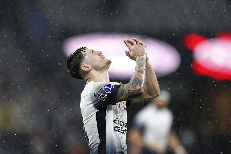 Um jogador de futebol está de pé, com as mãos unidas em oração, olhando para cima. Ele está molhado devido à chuva, e seu uniforme é predominantemente branco com detalhes em preto. O fundo é desfocado, com luzes e gotas de água visíveis no ar, sugerindo um ambiente de jogo intenso.