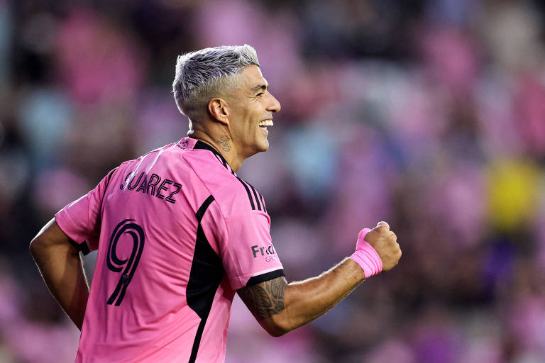 Um jogador de futebol está comemorando, vestindo uma camisa rosa com o nome 'SUAREZ' e o número 9 nas costas. Ele tem cabelo platinado e está sorrindo, levantando o punho em sinal de celebração. O fundo da imagem é desfocado, com tons de rosa, sugerindo um ambiente de jogo animado.
