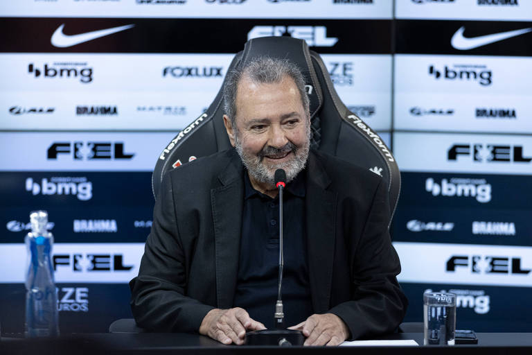 Um homem sorridente está sentado em uma mesa de coletiva de imprensa, com um microfone à sua frente. Ele usa uma camisa preta e um blazer escuro. Ao lado dele, há uma garrafa de água e um copo. O fundo é composto por painéis com logotipos de patrocinadores, incluindo Nike e BMG.
