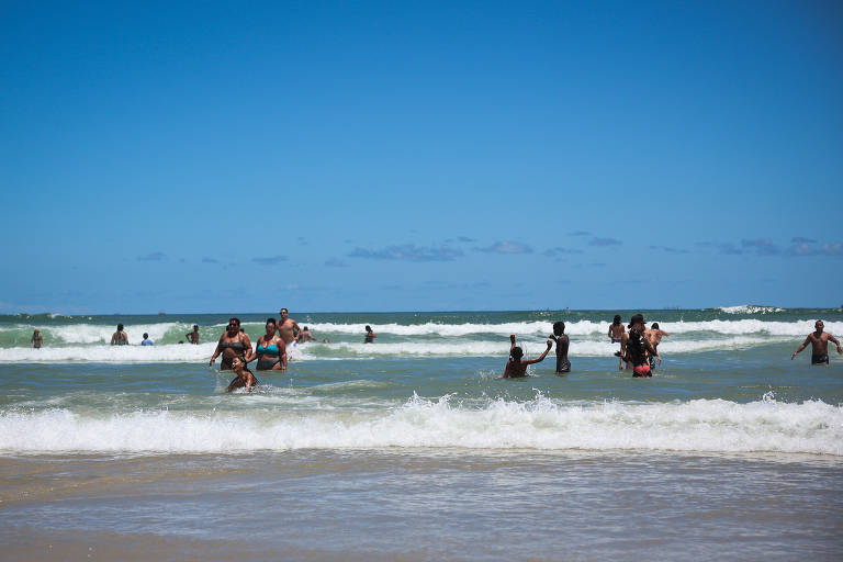 A imagem mostra várias pessoas se divertindo na água do mar em uma praia. Algumas estão de pé nas ondas, enquanto outras estão parcialmente submersas. O céu está claro e azul, com algumas nuvens esparsas. As ondas são visíveis, e a areia da praia é clara e seca.