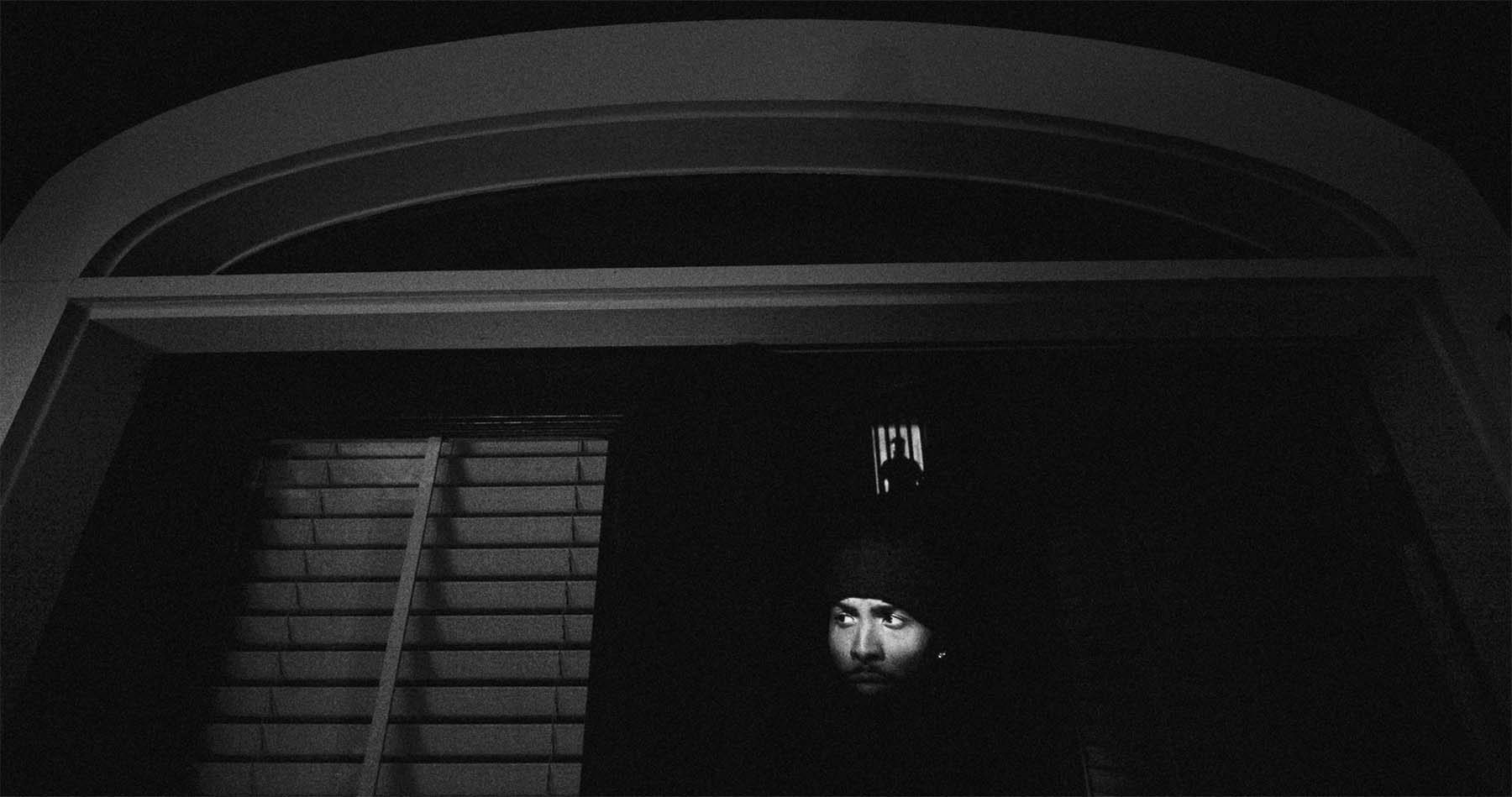 Man wearing a beanie looks out his front door into the dark. A creepy figure stands in a lit doorway in the background.