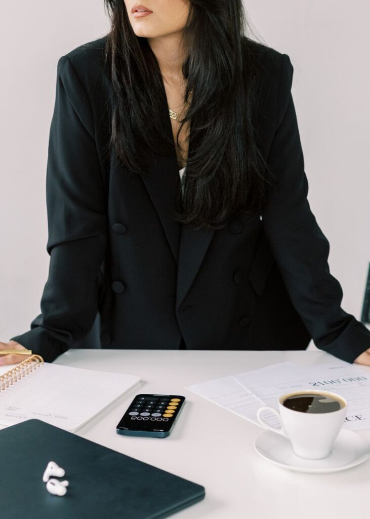 image showing a business woman in blazers with hands on a table while standing. A cup of coffee, calculator and laptop on her desk