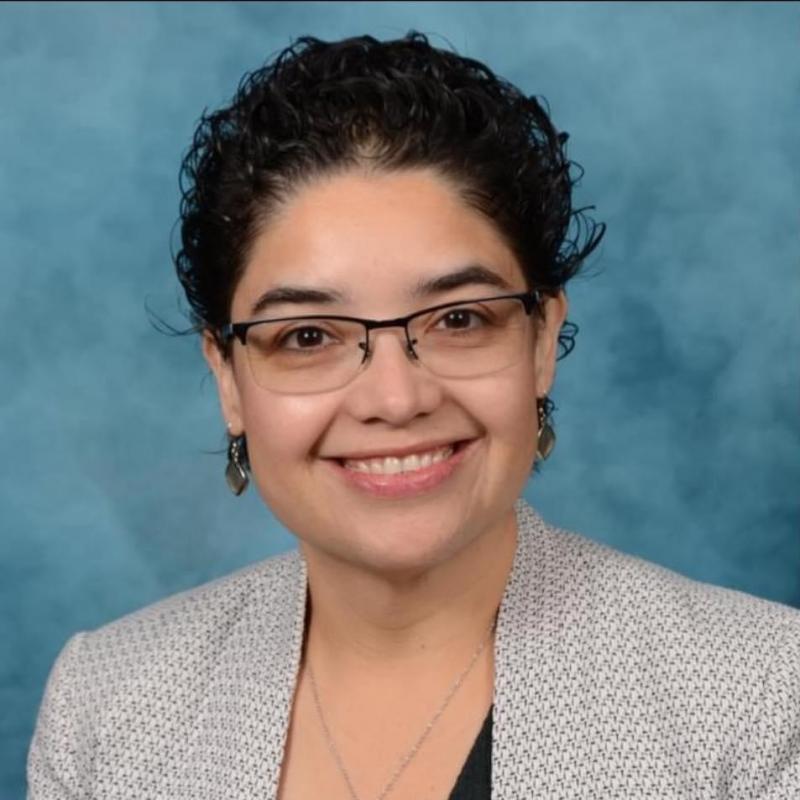 A woman with short, curly hair smiles warmly at the camera, wearing glasses and a patterned blazer.