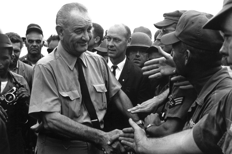 Lyndon B. Johnson is surrounded by soldiers in this black-and-white photo as he visits U.S. troops in Vietnam. Johnson smiles as he shakes hands with a service member wearing uniform. He is flanked by security officers in dark suits.