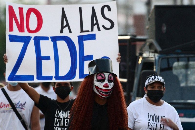 A person with a top hat, long red hair, and evil clown make-up stands in the central foreground as a man in the near background holds up a white protest sign reading "No A Las ZEDEs", translated to "No to the Employment and Development Zones".