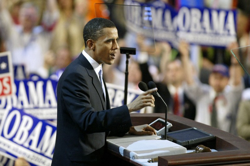 Barack Obama stands at a lectern with blurred faces and signs behind him.