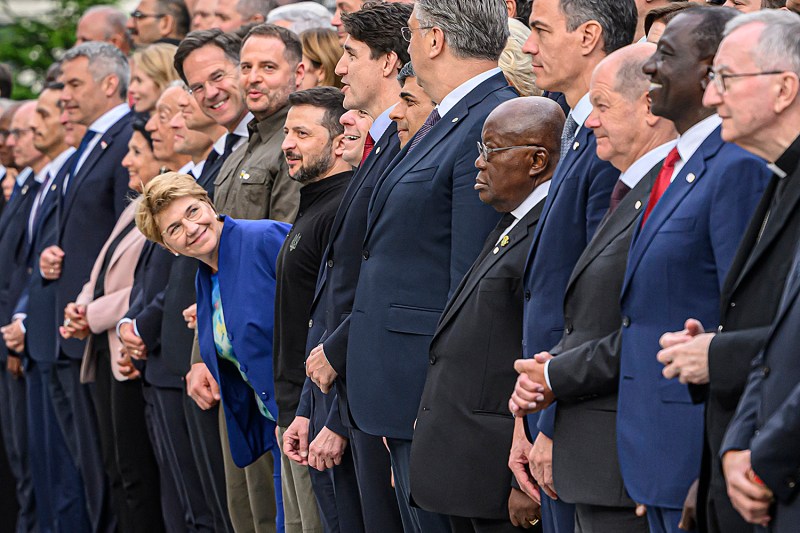 A woman in a blue suit leans forward to look down the line of world leaders and dignitaries, many in suits.