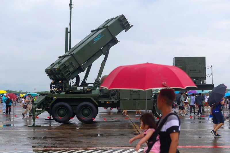 A man wearing a baby in a carrier holds an umbrella as he walks by a missile system. Other people mill about in the distance.