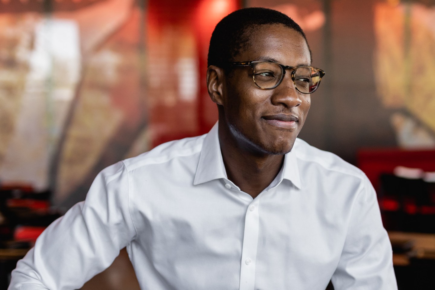 Damola Adamolekun wear a white button up and glasses and smiles while looking to his left.