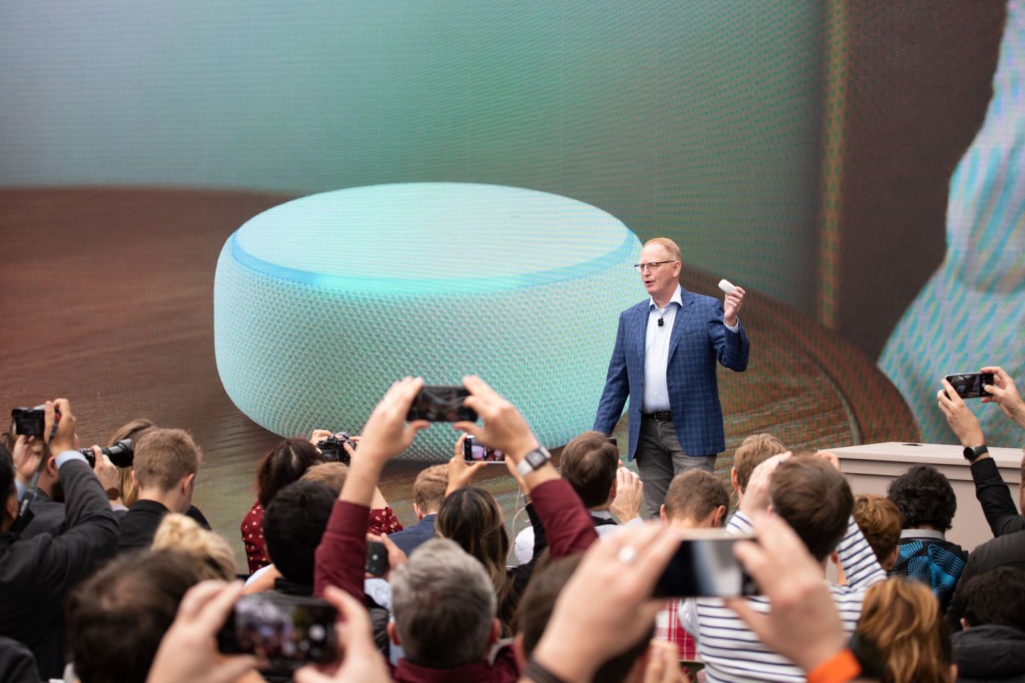 A man in a blue suit holds up a small circular device in front of a screen with the same device projected onto it. People in front of the stage hold up phones, taking pictures.