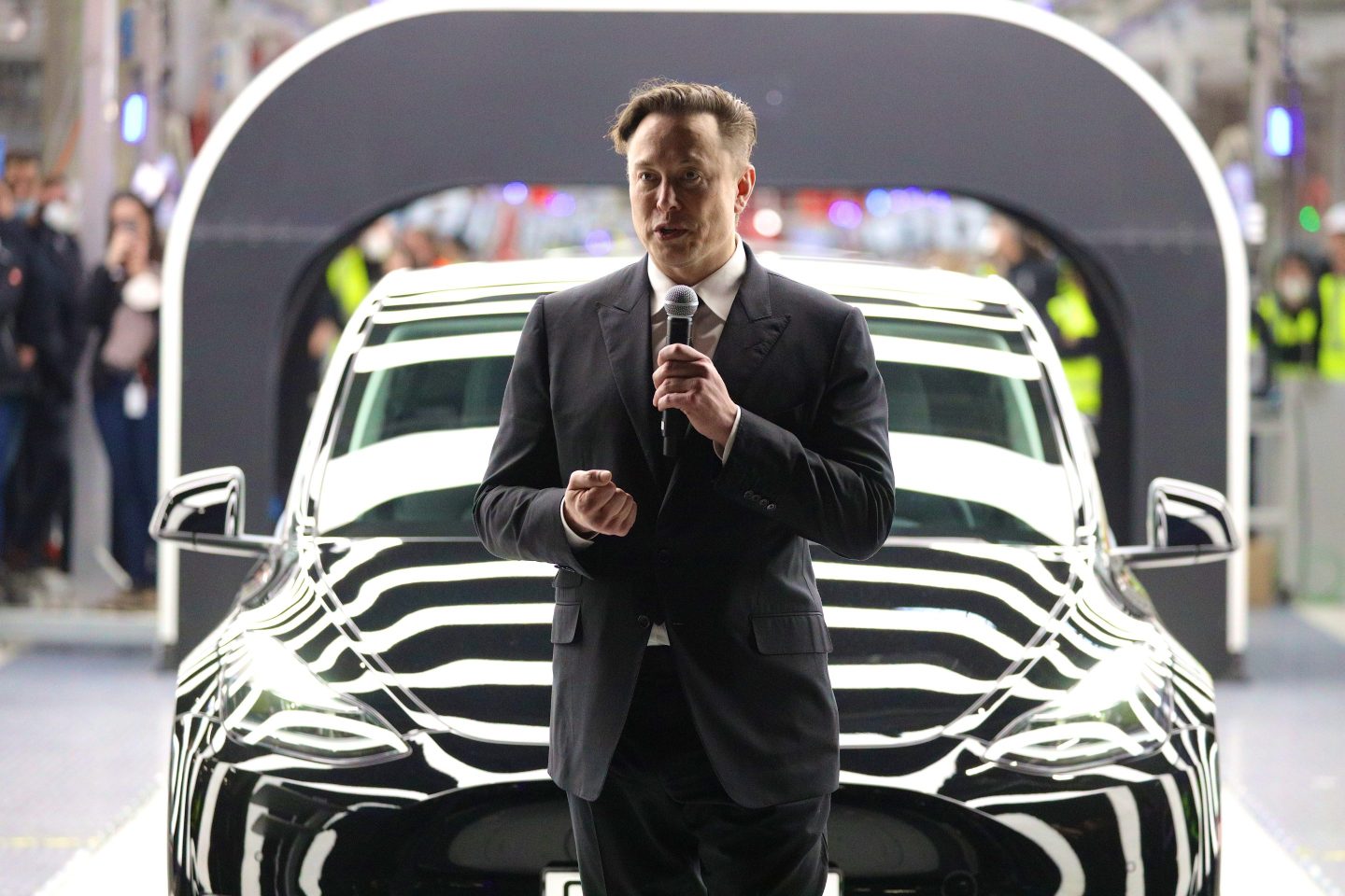 Elon Musk stands with a microphone in front of a lit up Tesla.