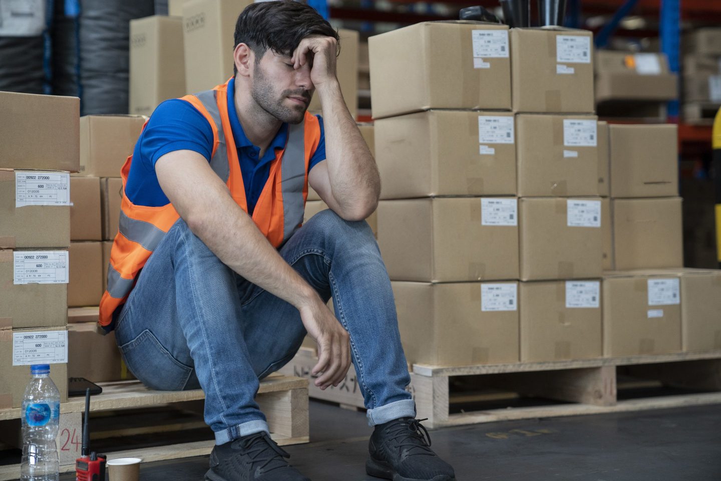 A man in an orange safety vest sits on a crate with his head in his hand, looking disappointed.