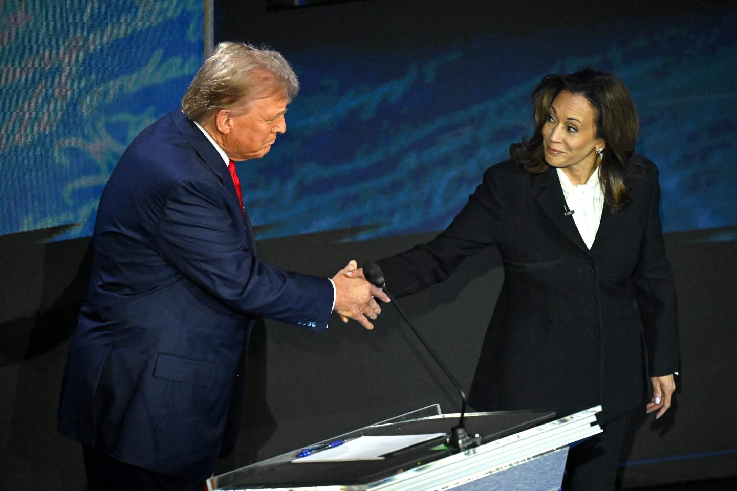 Former President Trump and Vice President Harris shake hands on the debate stage near Trump's podium. Both are wearing dark suits.