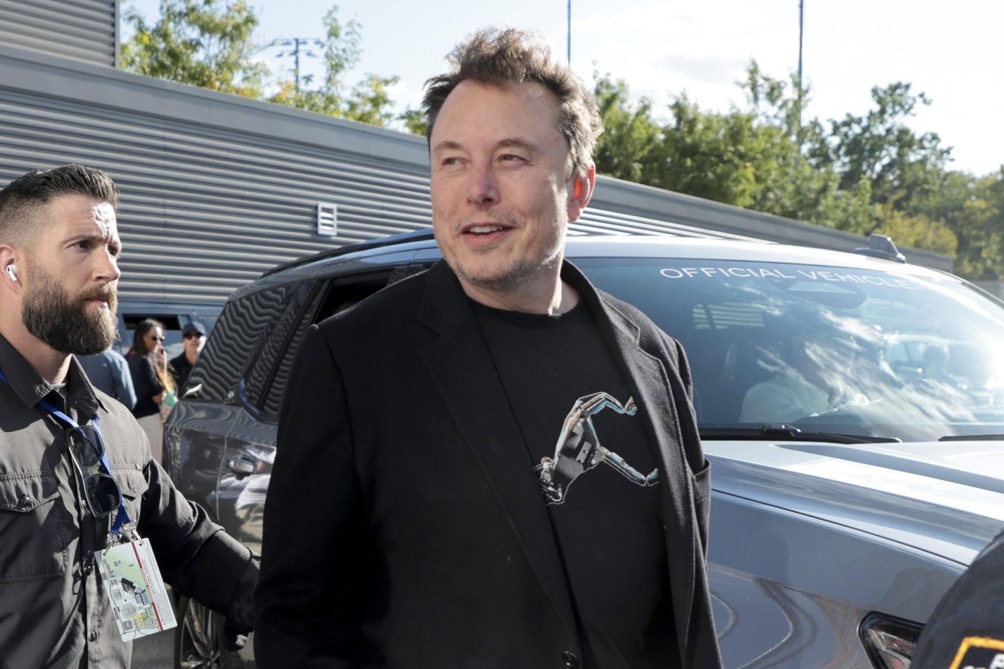 Elon Musk, dressed in a black t-shirt and blazer, looks to the left of the image and smiles as he's escorted by security.