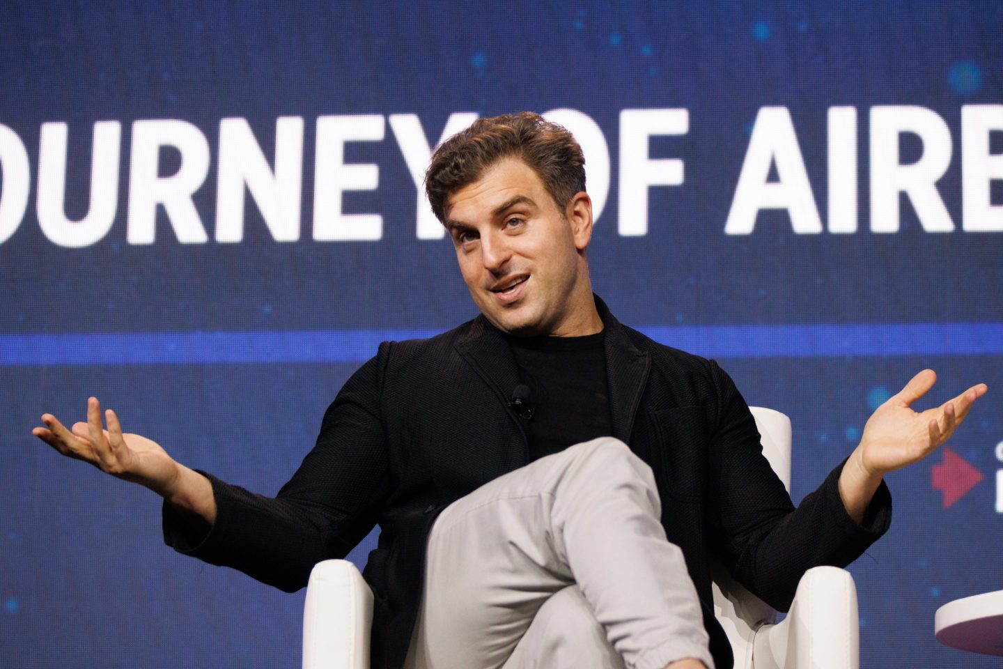 Brian Chesky sits in a chair on stage with his legs crossed and holding his arms up.