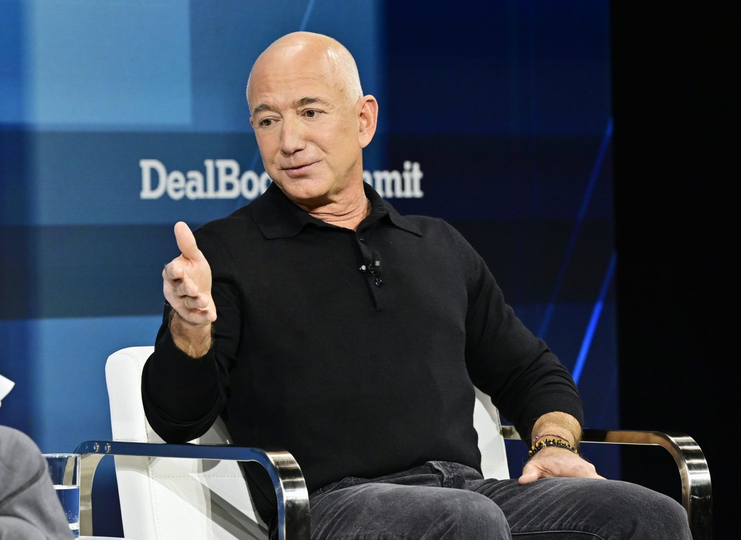 Jeff Bezos sits in a chair with his hand up looking away from the camera.