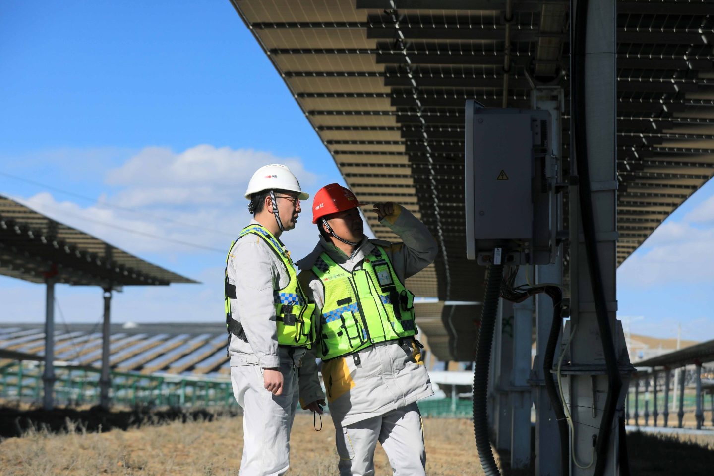 Two men in helmets and bright green vests inspect the underbelly of a solar panel.