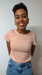 Imagem de Beatriz de Oliveira. Ela aparece sorrindo. Beatriz tem cabelos curtos e usa iuma camiseta cor-de-rosa e calças jeans.