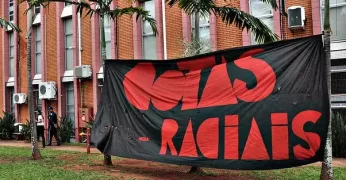 A imagem mostra um dos prédios da Unicamp. Há uma faixa preta, com fontes vermelhas, com a frase "Cotas raciais". A imagem ilustra a nota sobre a Lei de Cotas.