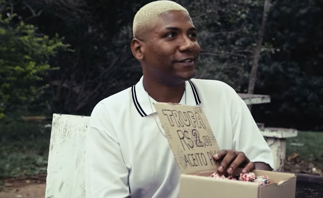 A imagem é uma cena do filme "Neurótico & Consciente" na qual Pedro, o protagonista, aparece segurando uma caixa de trufas. Ele veste uma camiseta branca, do Santos Futebol Clube, e tem cabelo em altura extremamente baixa e pintado em tons claros.