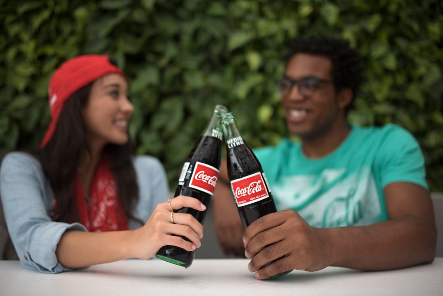 Two people clanking their Coke bottles together while seated and chatting outside. 