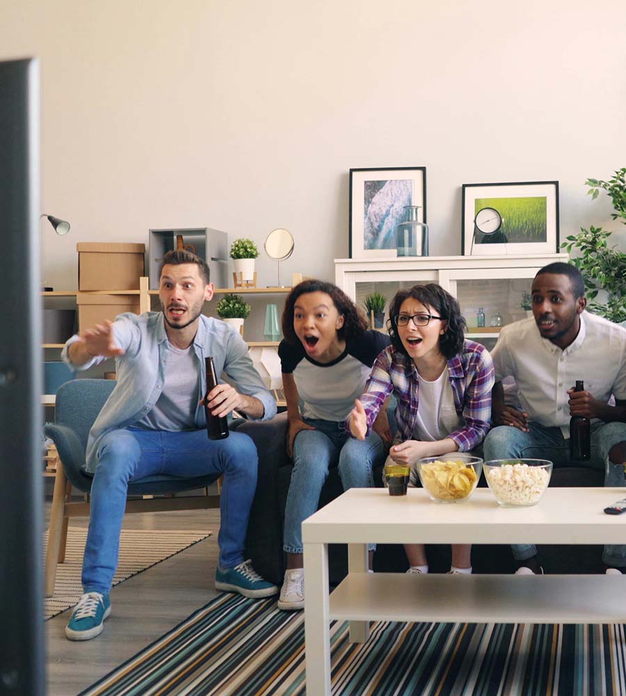 Four friends enthusiastically watch a sports game on TV, seated on a couch with snacks like popcorn and chips on the table. They react with excitement, possibly witnessing a pivotal moment in a women's basketball game, as women's sports continue to gain significant attention and support from fans and sponsors alike.