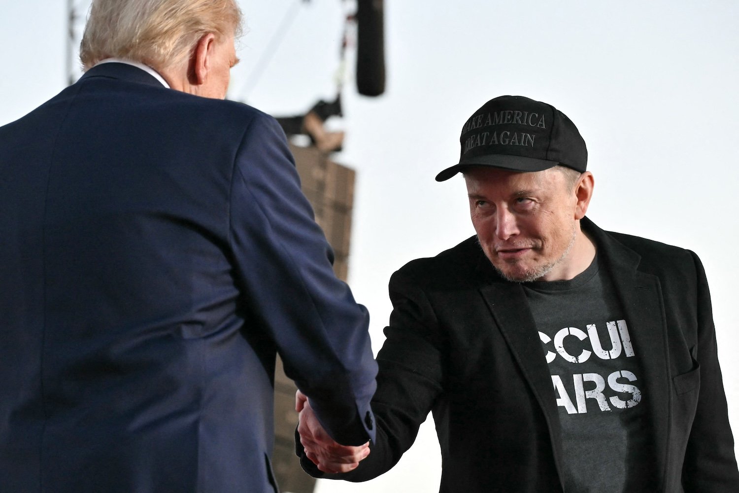 Elon Musk shaking the hand of Donald Trump in the foreground, back to the camera.