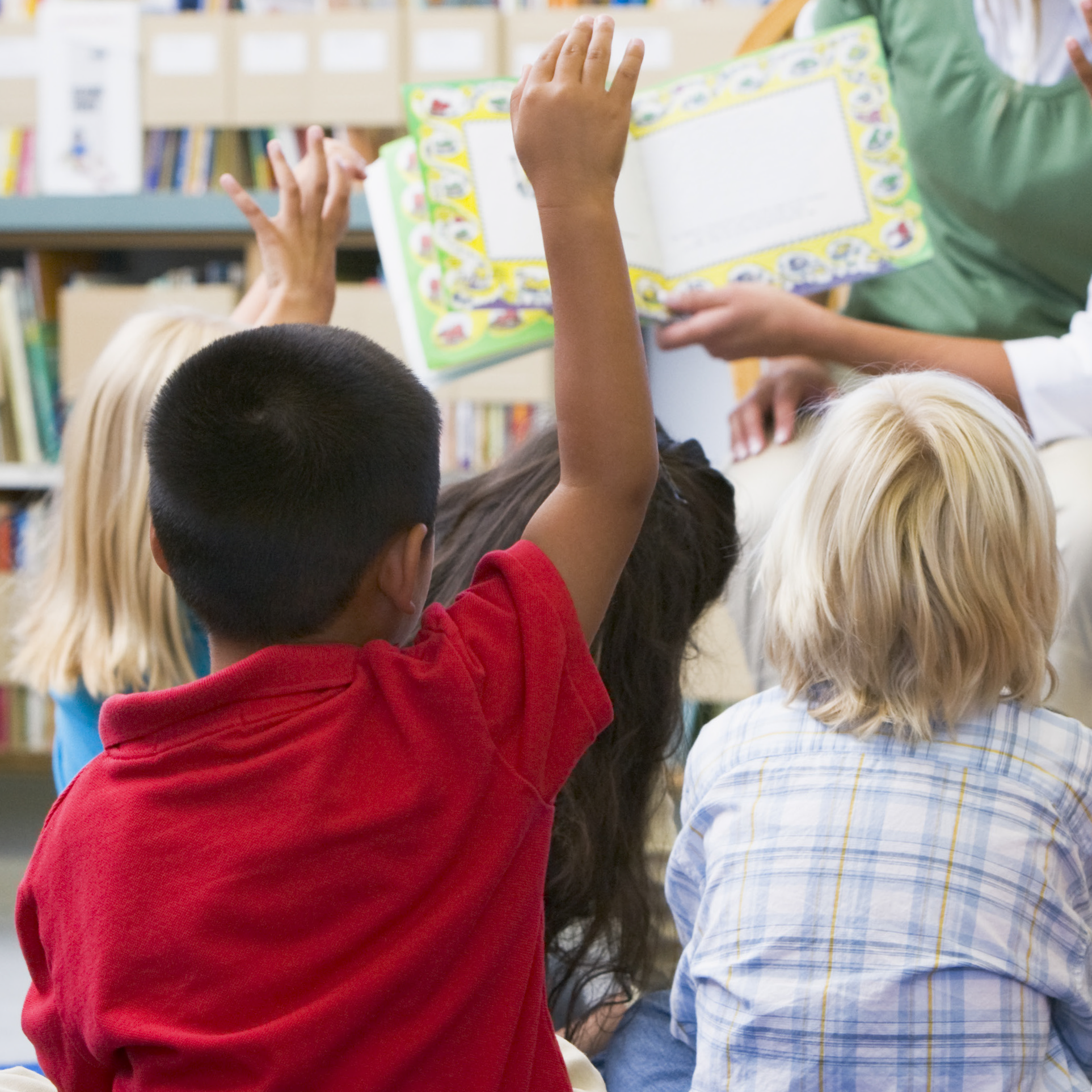 Children sit in a classroom, raising their hands and listening to a teacher read them a book