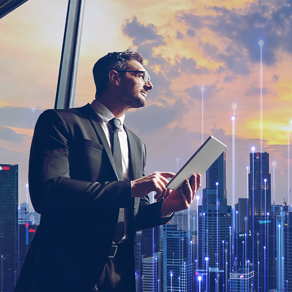 A businessman holds a tablet and stares out the office window at city buildings