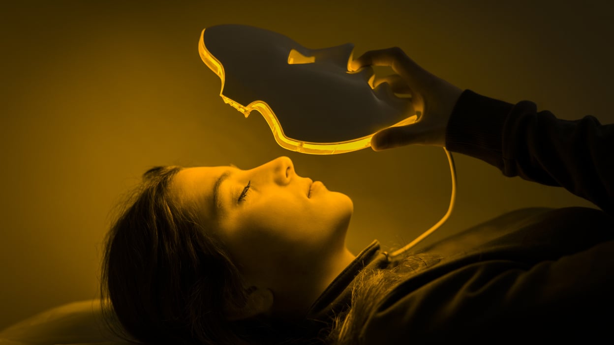 Woman laying down is putting on a face mask with eye, nose, and mouth cutouts. The mask is lit with yellow colored lights from within. 