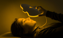 Woman laying down is putting on a face mask with eye, nose, and mouth cutouts. The mask is lit with yellow colored lights from within. 