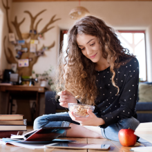 Female student sitting on floor, reading books, studying for final exam at university. Beautiful student at home eating cereals.