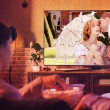 A woman watches a period drama TV show from her couch, probably set in the 19th century.