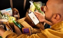 a person looks into several bags full of groceries while holding a cell phone in their right hand that displays the carrot logo for instacart