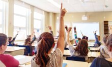 Several children sitting in a classroom, photographed from behind, eagerly raise their hands.