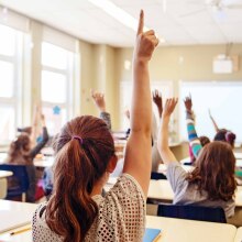 Several children sitting in a classroom, photographed from behind, eagerly raise their hands.